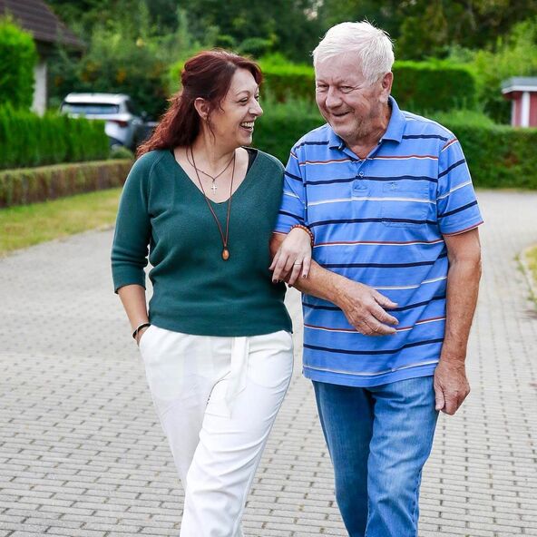 Eine lächelnde Betreuerin geht mit einem Demenzkranken über einen gepflasterten Weg in einer grünen Umgebung.