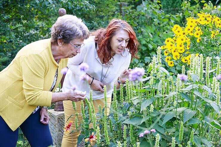 Eine Seniorenbetreuung findet im Garten statt, wo eine Demenzerkrankte und eine Helferin gemeinsam Pflanzen betrachten.
