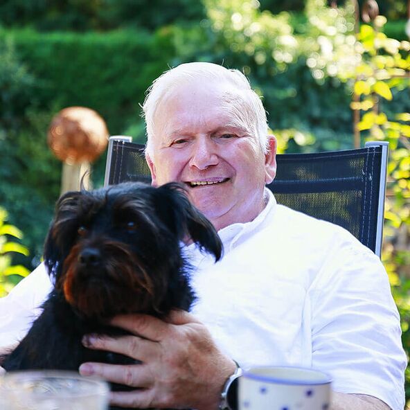 Ein lächelnder Senior mit einem Hund in den Armen genießt die Zeit im Freien, während er liebevolle Betreuung erhält.