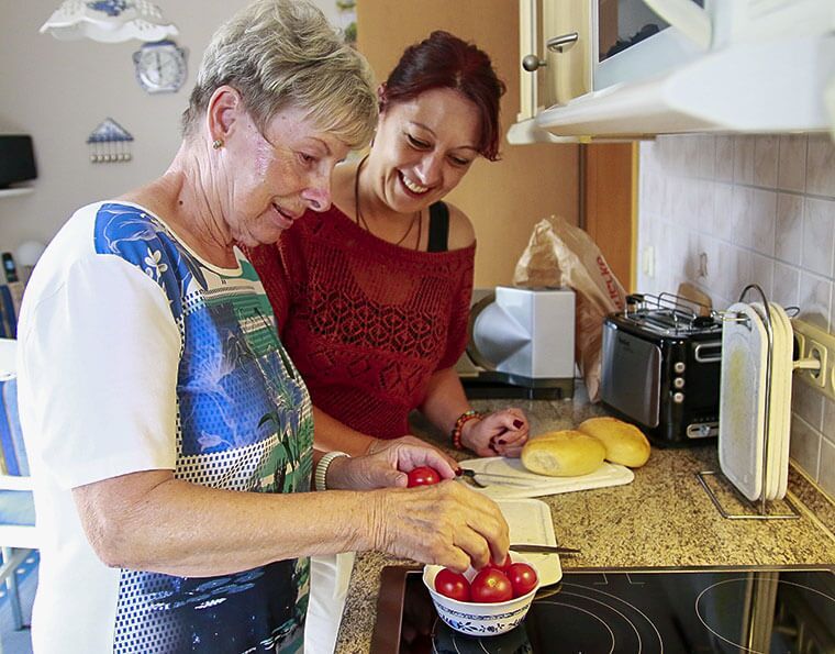 Eine Seniorin und ihre Betreuerin bereiten gemeinsam frische Tomaten in der Küche vor, um eine Mahlzeit zuzubereiten.