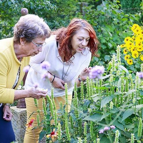Eine Seniorenbetreuung findet im Garten statt, wo eine Demenzerkrankte und eine Helferin gemeinsam Pflanzen betrachten.